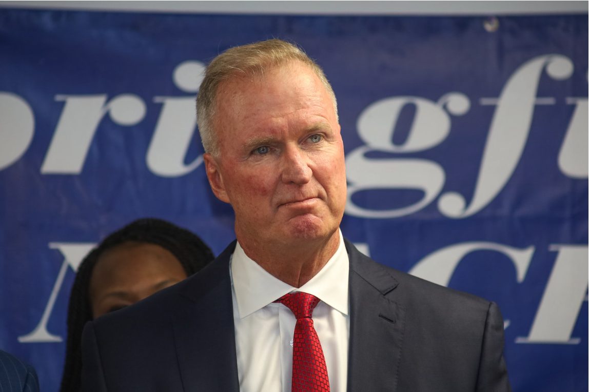 Man with short white hair, wearing a dark blue suit, which dress shirt, and a red tie. He stands in front of the NAACP Springfield sign.