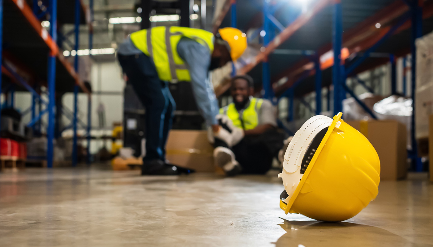 At the forefront of the image is a yellow hard hat, in the background is a man, wearing bright yellow safety vest and yellow hard hat. There is another man on the ground, indicating an industrial accident.